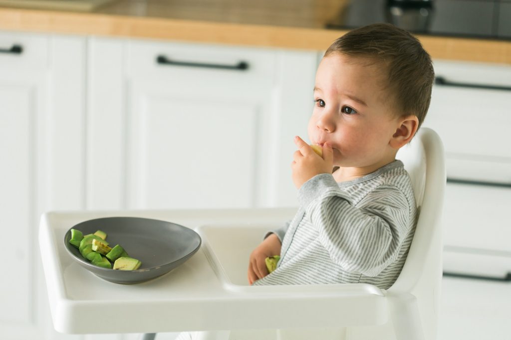 Happy baby sitting in high chair eating fruit in kitchen Healthy nutrition for kids Bio carrot as - SKIDamarink KIDs Happy baby sitting in high chair eating fruit in kitchen. Healthy nutrition for kids. Bio carrot as