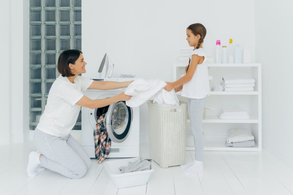 Happy mother loads clothes in washing machine, little girl helps, gives white linen from basket