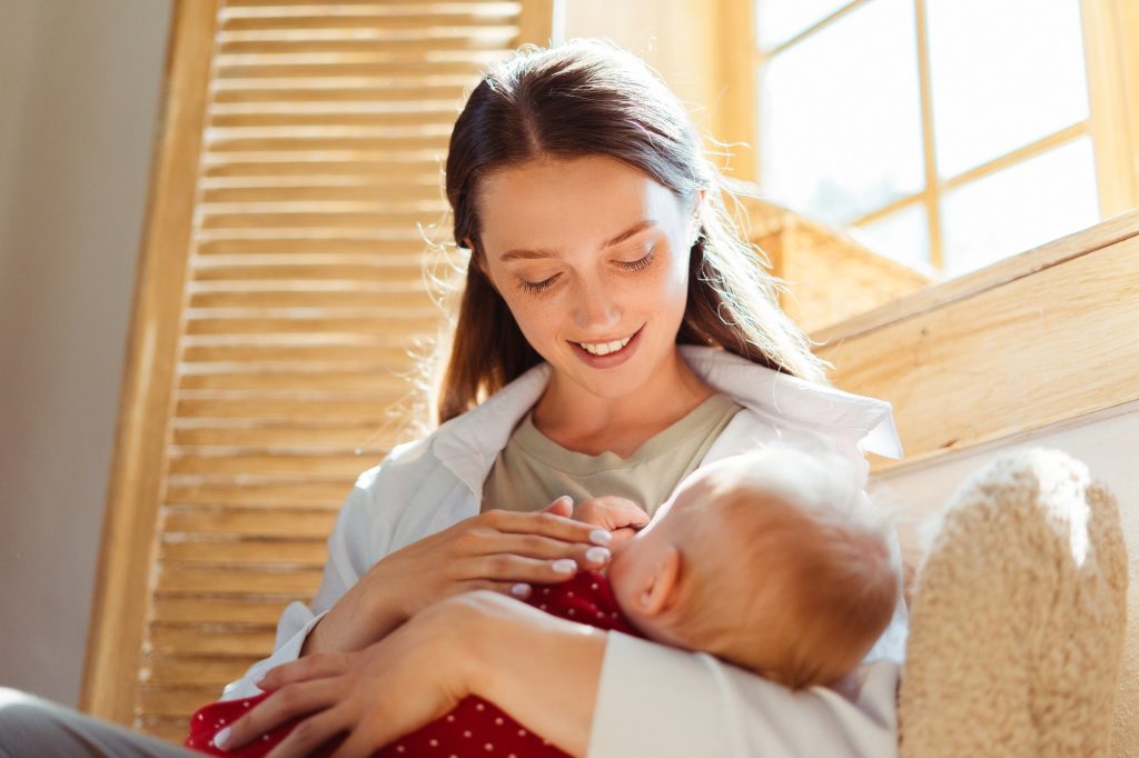 Smiling mother holding her little cute baby breastfeeding at home Concept of breastfeeding - SKIDamarink KIDs Smiling mother holding her little cute baby breastfeeding at home. Concept of breastfeeding