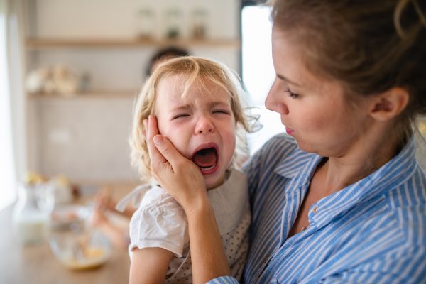 A mother holding a crying toddler daughter indoors in kitchen - SKIDamarink KIDs A mother holding a crying toddler daughter indoors in kitchen.