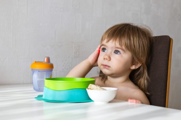 A young toddler girl with curly hair dressed in pink overalls sits at a wooden table - SKIDamarink KIDs A young toddler girl with curly hair, dressed in pink overalls, sits at a wooden table.