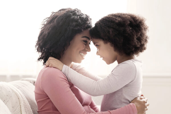 Mother and daughter share a warm hug, smiling with foreheads together in soft light.